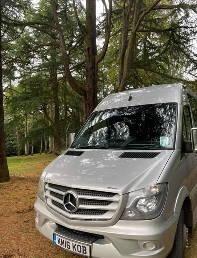Front of a silver Mawley Minibus Mercedes Sprinter under tall trees